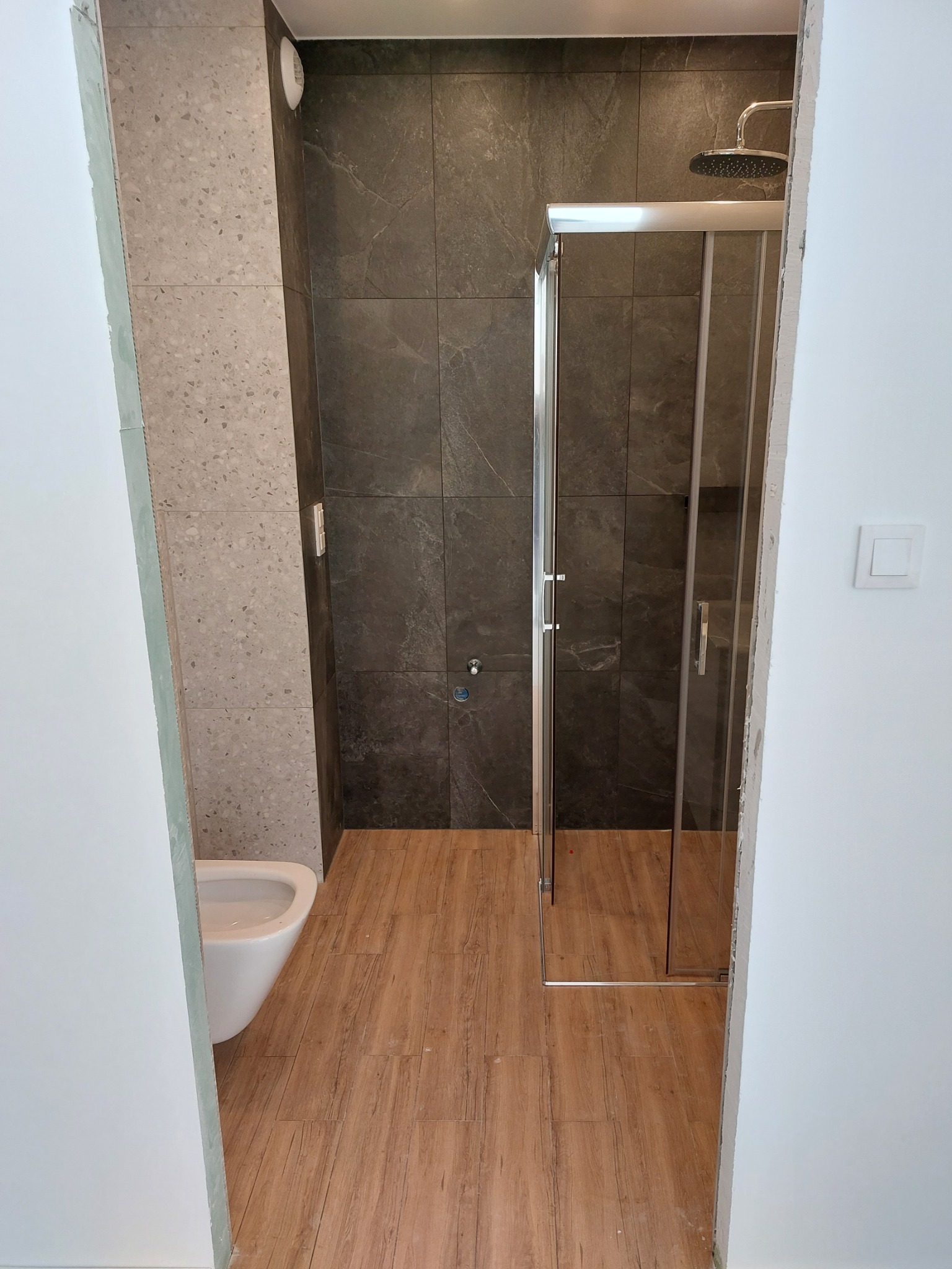 Wykończona łazienka z kabiną prysznicową ze srebrnymi ramami, ciemnymi płytkami imitującymi kamień na ścianach prysznica, toaletą wiszącą i podłogą z paneli imitujących drewno.