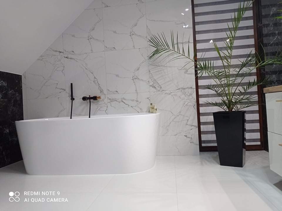 Nowoczesna łazienka z wolnostojącą białą wanną, marmurowe płytki na ścianach, czarna armatura i wysoka roślina w czarnej donicy, żaluzje poziome w oknie.