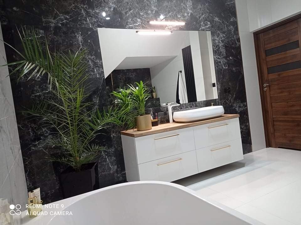 Nowoczesna łazienka z czarną marmurową ścianą, białą szafką pod umywalkę z drewnianym blatem, dużym lustrem i roślinami doniczkowymi, częściowo widoczna biała wanna na pierwszym planie.