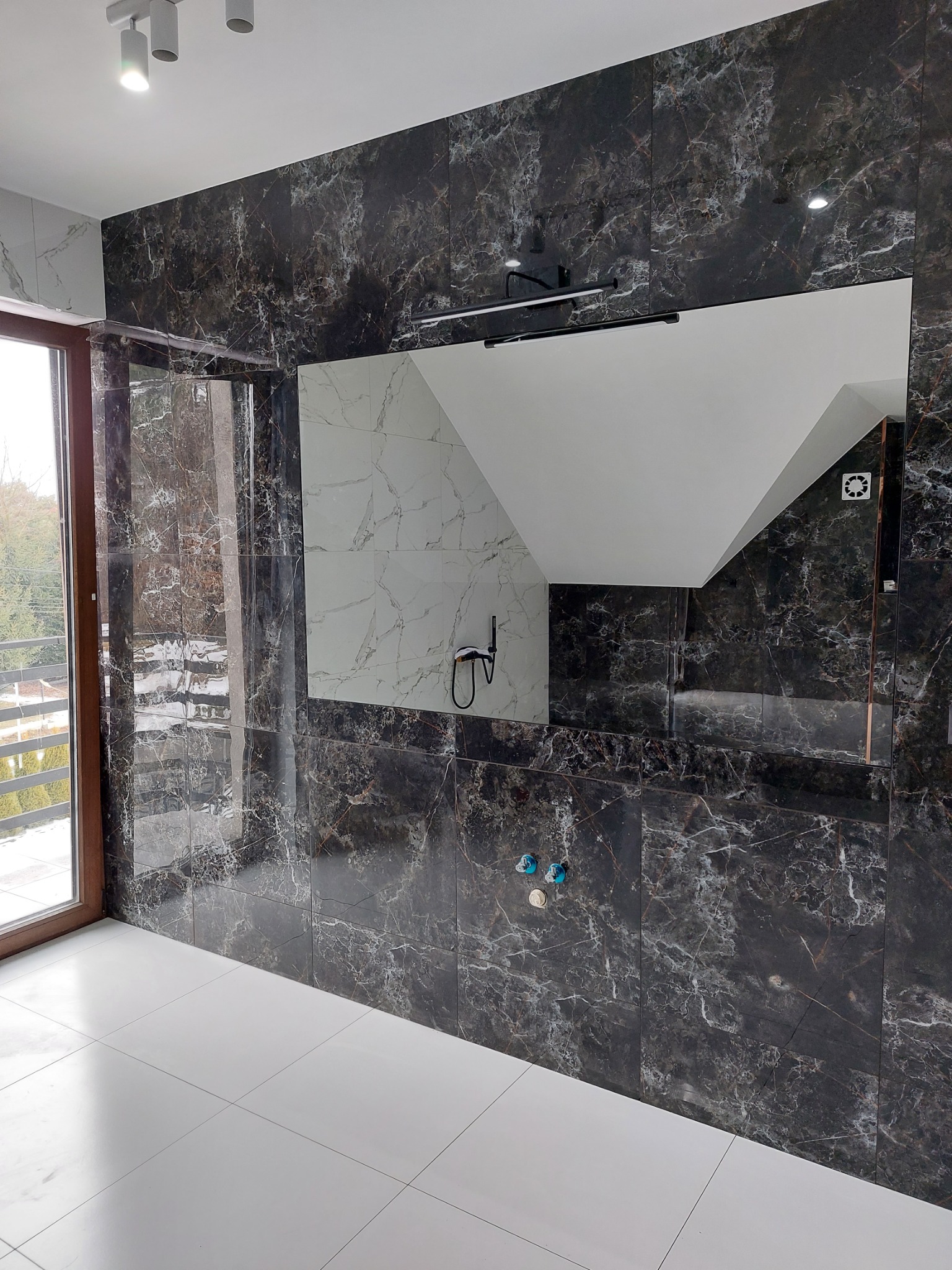 Elegancka łazienka z czarnymi płytkami imitującymi marmur na ścianach, białymi płytkami na podłodze, dużym lustrem z oświetleniem i widokiem z okna na zewnątrz.