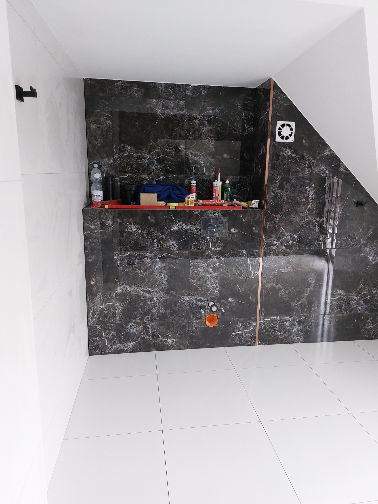 Wnętrze łazienki w trakcie remontu, z ciemnymi płytkami imitującymi marmur na ścianach i białymi płytkami na podłodze; widoczny fragment skośnego sufitu, wentylator, butelka wody i narzędzia...