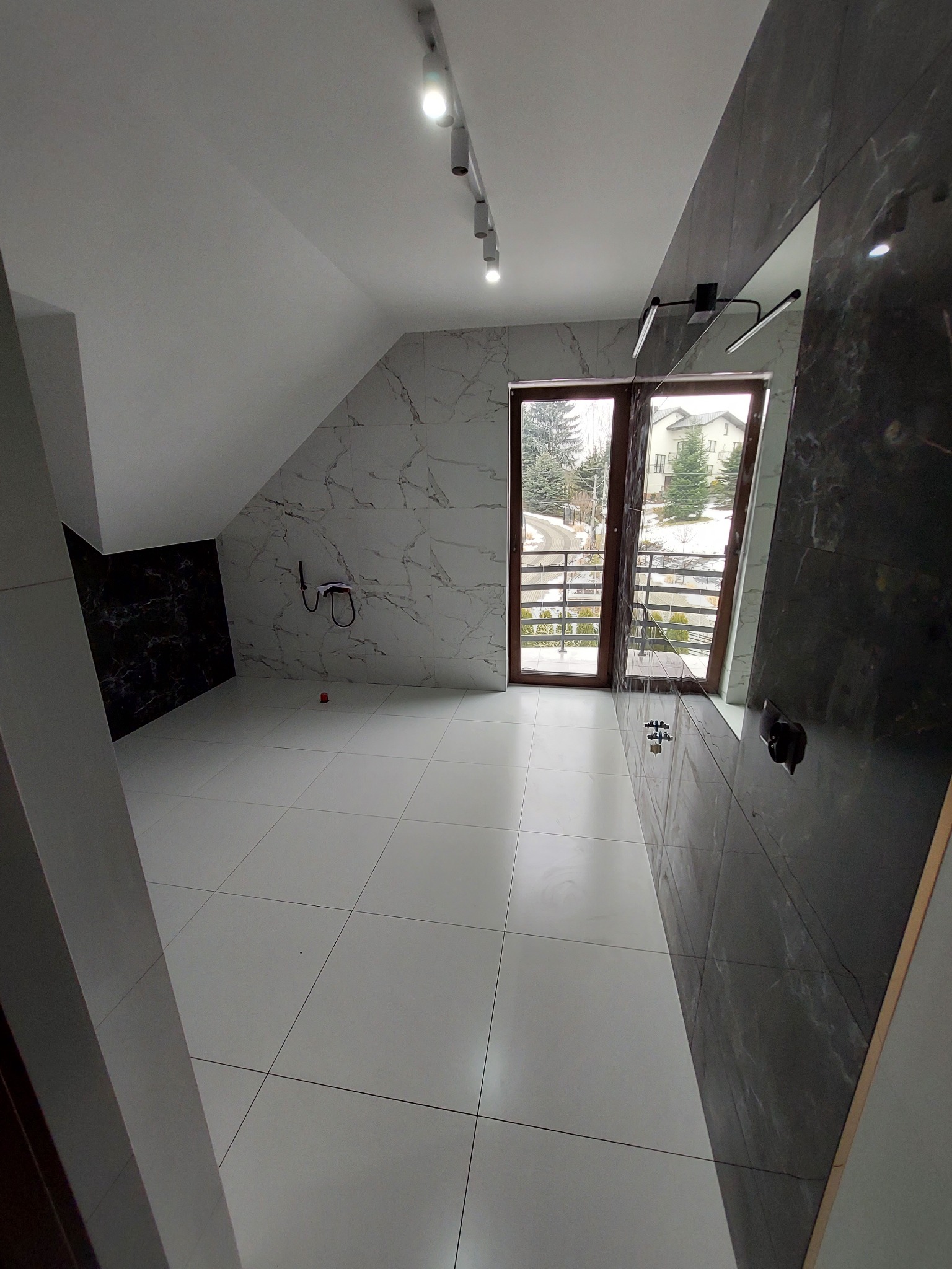 Jasna łazienka na poddaszu z oknem balkonowym, białe płytki podłogowe i ścienne imitujące marmur, czarna ściana z instalacją prysznicową i lustrem, oświetlenie punktowe na suficie.