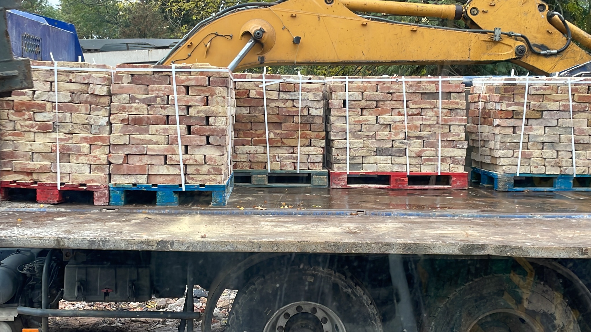 Cegły klinkierowe ułożone na paletach w kolorach czerwonym i niebieskim, załadowane na platformę ciężarówki z ramieniem koparki w tle.