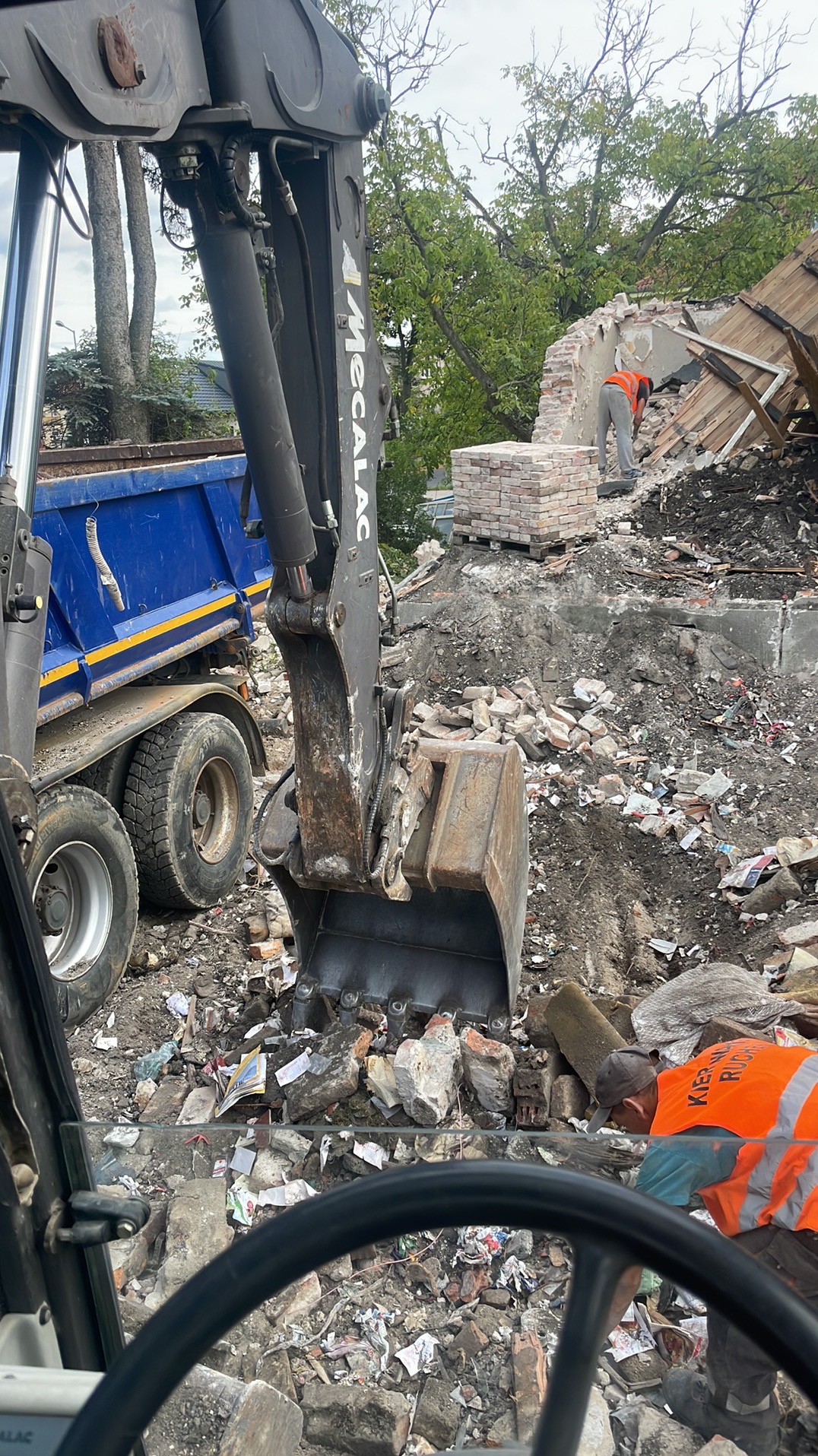Widok z kabiny operatora na plac budowy z koparką Mecalac, gruzem i pracownikiem w pomarańczowej kamizelce, demontującym pozostałości muru.