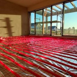 Hydrololo Karol Kleisa - Rozłożone, czerwone rurki ogrzewania podłogowego na srebrnej folii izolacyjnej w surowym wnętrzu z dużym oknem tarasowym i widokiem na budynki w oddali.