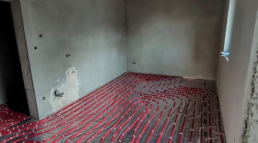 Czerwone rury ogrzewania podłogowego ułożone na siatce w surowym pomieszczeniu z widocznymi niedoskonałościami tynku i małymi otworami w ścianie.