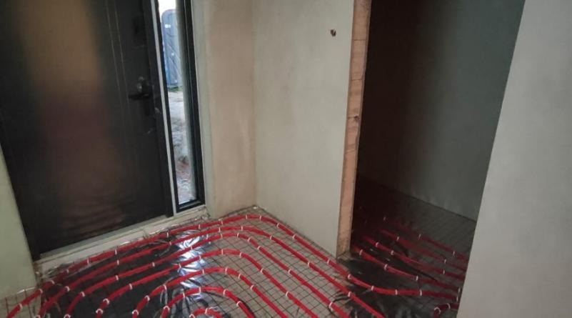 Czerwone rury ogrzewania podłogowego ułożone na srebrnej folii izolacyjnej w surowym pomieszczeniu z otynkowanymi ścianami i widocznym progiem drzwiowym.