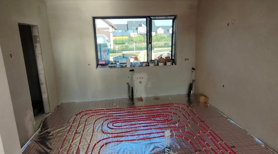 Wnętrze w stanie deweloperskim z rozłożonym systemem ogrzewania podłogowego na folii aluminiowej, widoczne rury w kolorze czerwonym ułożone w pętle, okno z widokiem na osiedle domów jednorodzinnych.