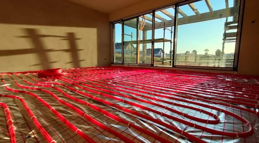 Rozłożone, czerwone rurki ogrzewania podłogowego na srebrnej folii izolacyjnej w surowym wnętrzu z dużym oknem tarasowym i widokiem na budynki w oddali.