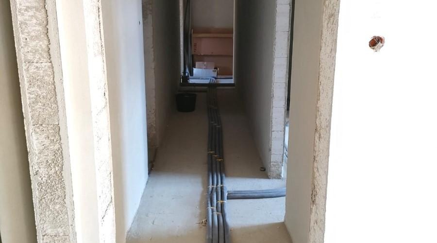 Korytarz w stanie surowym z ułożonymi rurami instalacyjnymi na podłodze, zabezpieczonymi taśmą, widoczne puszki elektryczne na ścianie.