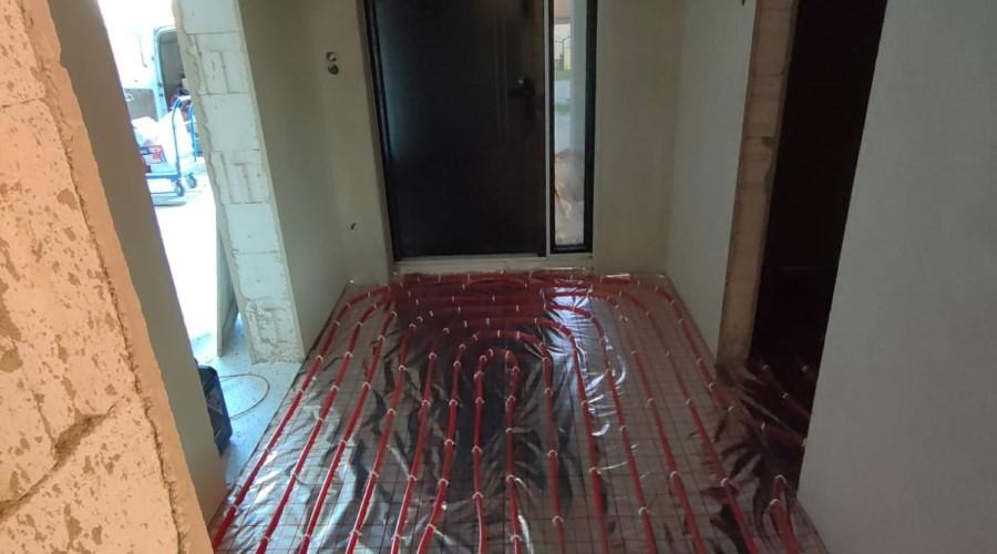 Czerwone rurki ogrzewania podłogowego rozłożone na srebrnej folii z siatką w surowym wnętrzu, widok na czarne drzwi wejściowe i fragment ściany z betonu komórkowego.