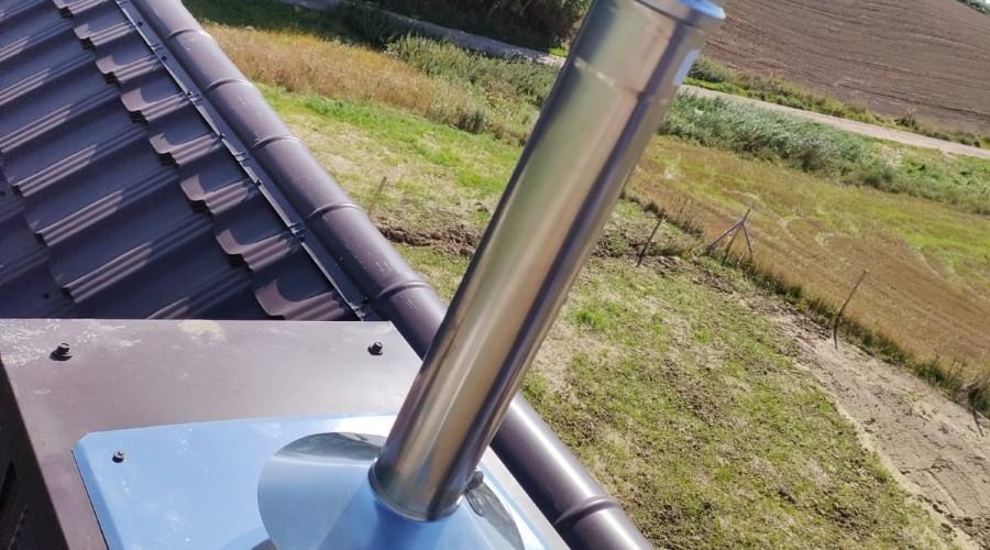 Błyszczący, metalowy komin wentylacyjny wystający z ciemnobrązowego dachu z dachówki ceramicznej na tle pola i nieba.