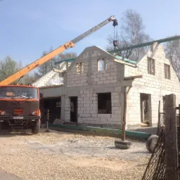 Wznoszenie domu z białych bloczków: surowa konstrukcja z otworami okiennymi, wspierana przez pomarańczowy dźwig. Widoczne elementy konstrukcyjne dachu.