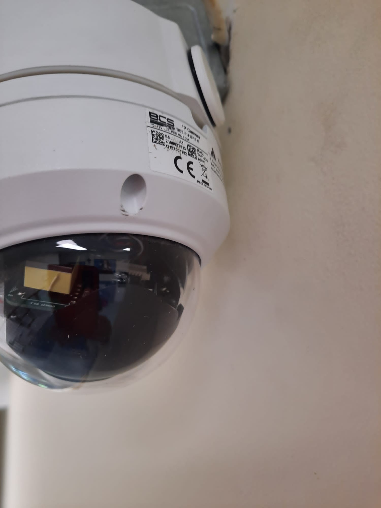 Biała kamera IP BCS zamontowana na ścianie, widoczne oznaczenia CE i numer seryjny.