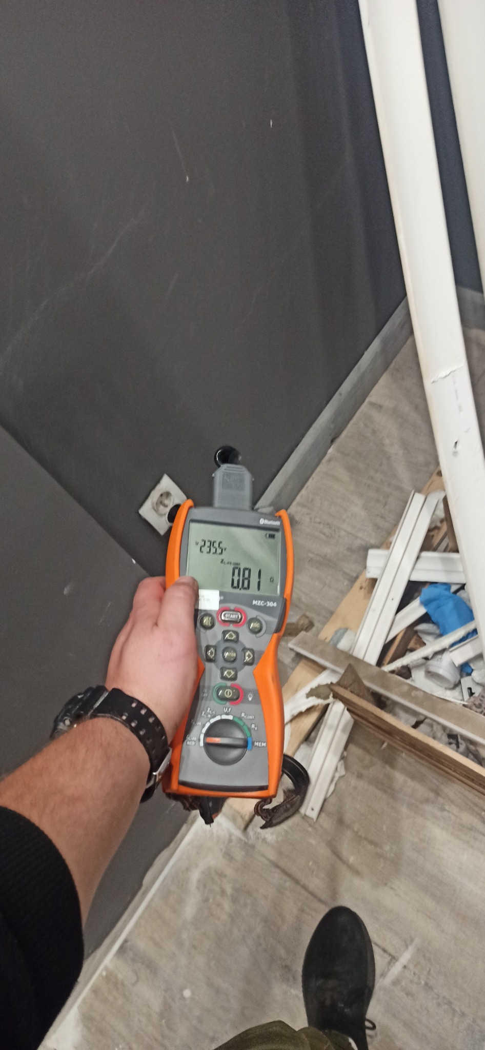 Elektryk mierzy parametry gniazdka elektrycznego za pomocą miernika MZC-304 na tle szarej ściany i podłogi w trakcie remontu.