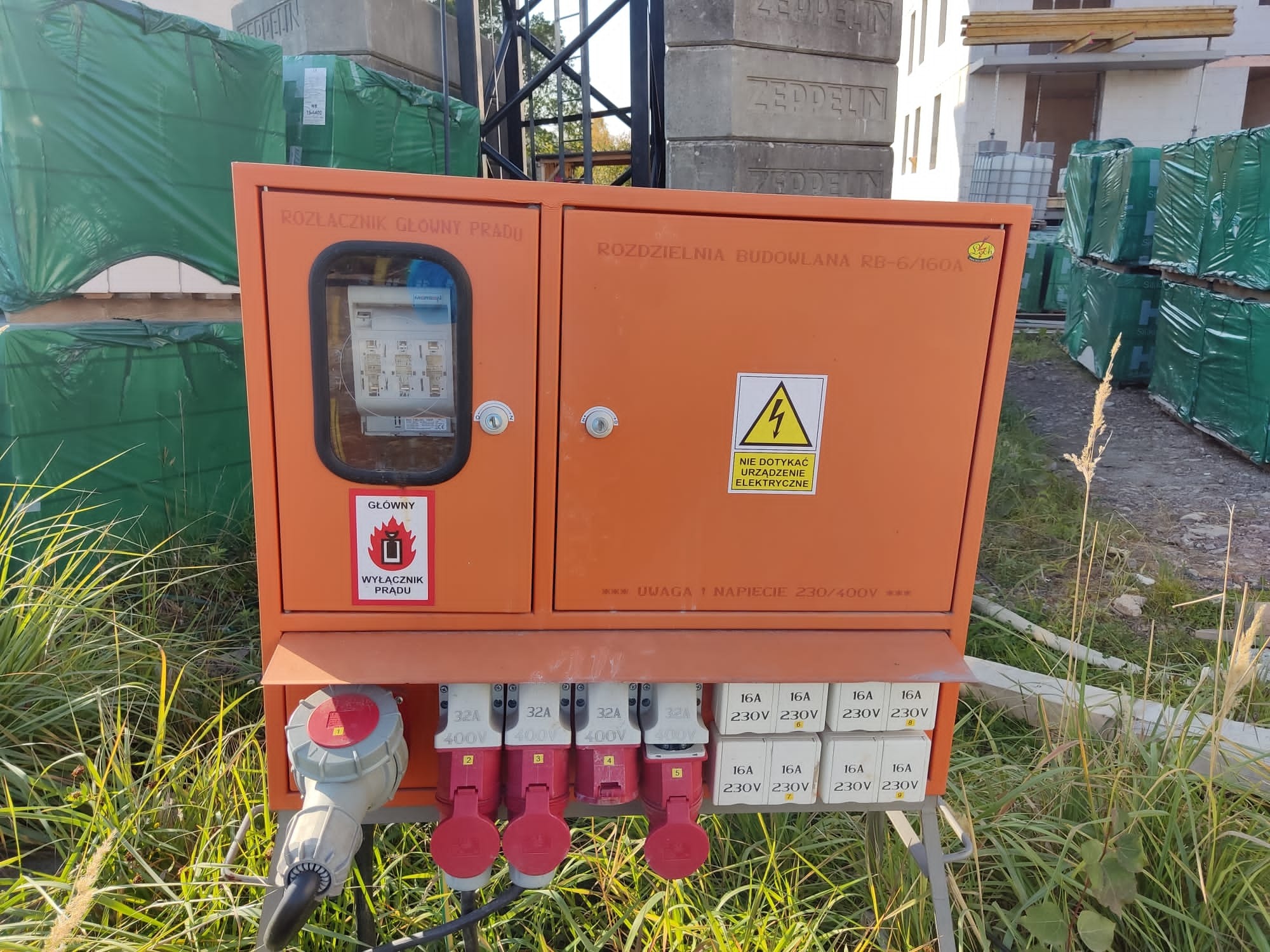 Pomarańczowa skrzynka rozdzielcza budowlana z widocznymi gniazdami 32A i 16A, etykietą ostrzegawczą i wyłącznikiem prądu, na tle materiałów budowlanych i trawy.