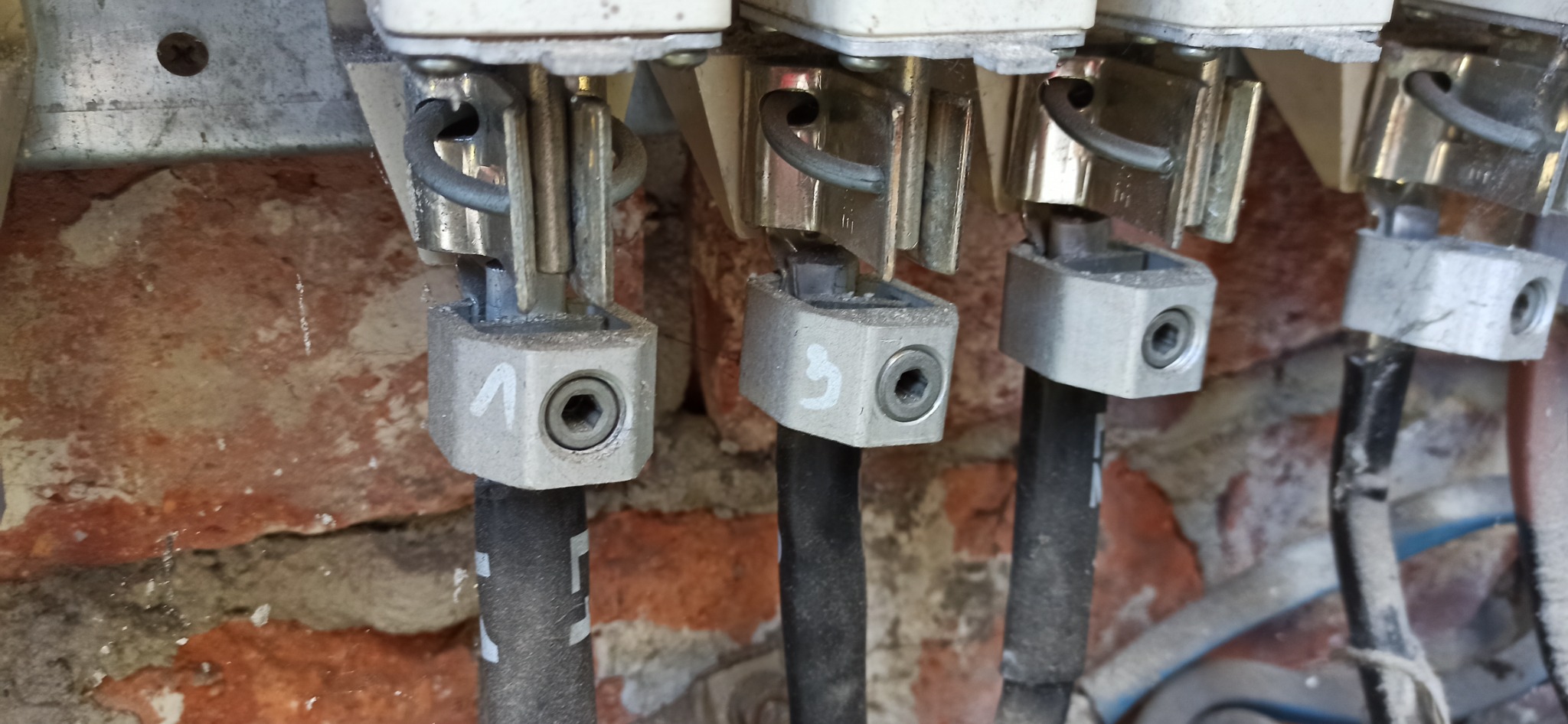 Szczegółowe ujęcie trzech przewodów elektrycznych przymocowanych do metalowych zacisków z numerami 1 i 3, na tle starej, częściowo otynkowanej ściany z czerwonej cegły.