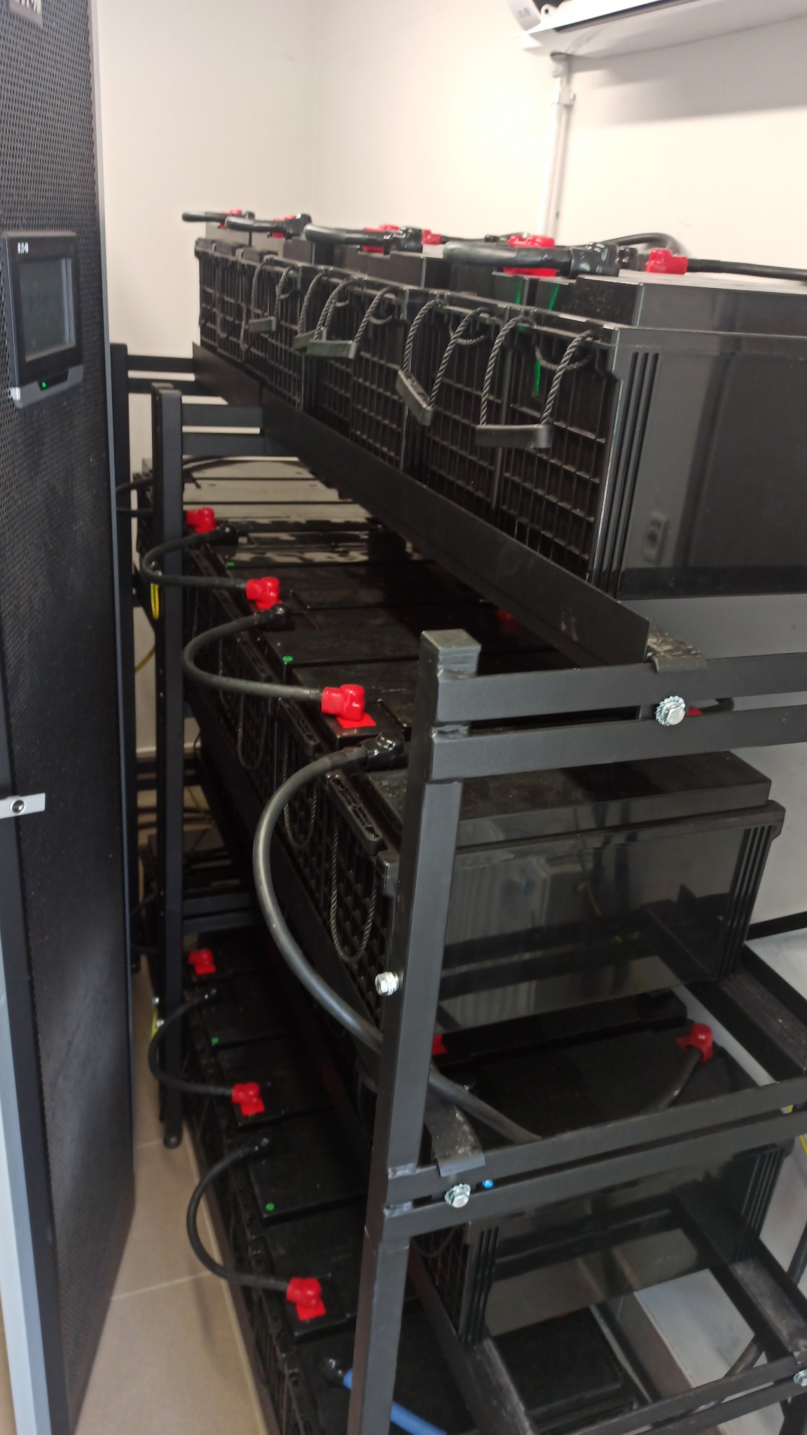 Baterie akumulatorowe na metalowym regale w pomieszczeniu technicznym, widoczne przewody z czerwonymi złączkami.