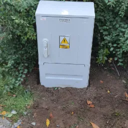 Szara skrzynka elektryczna INCOBEX z ostrzeżeniem o wysokim napięciu, umieszczona na ziemi obok zielonego krzewu.