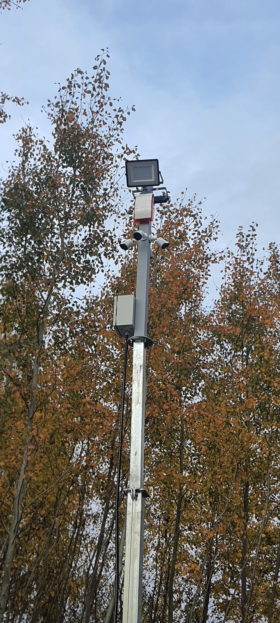 Słup monitoringu z kamerami i reflektorem LED na tle drzew z jesiennymi liśćmi i pochmurnego nieba.