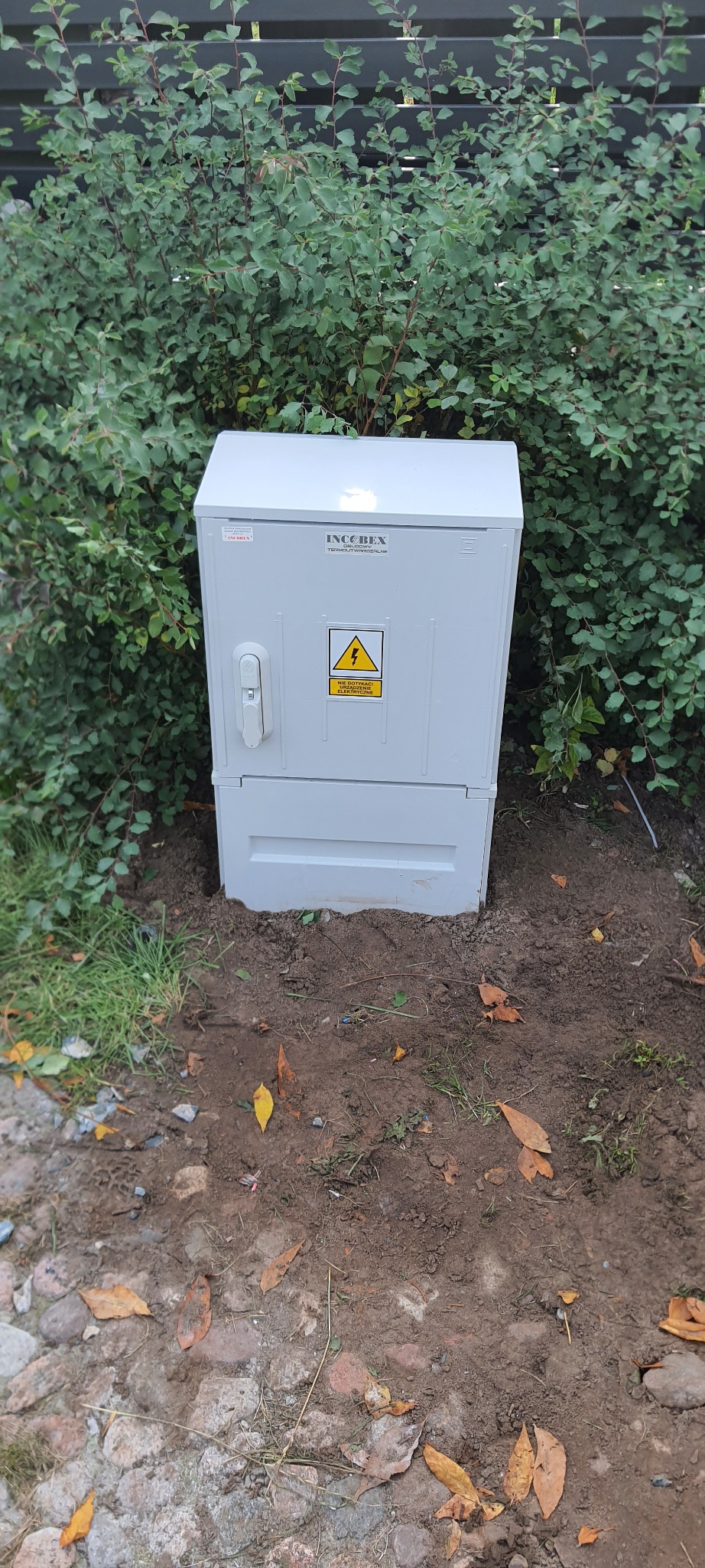 Szara skrzynka elektryczna INCOBEX z ostrzeżeniem o wysokim napięciu, umieszczona na ziemi obok zielonego krzewu.