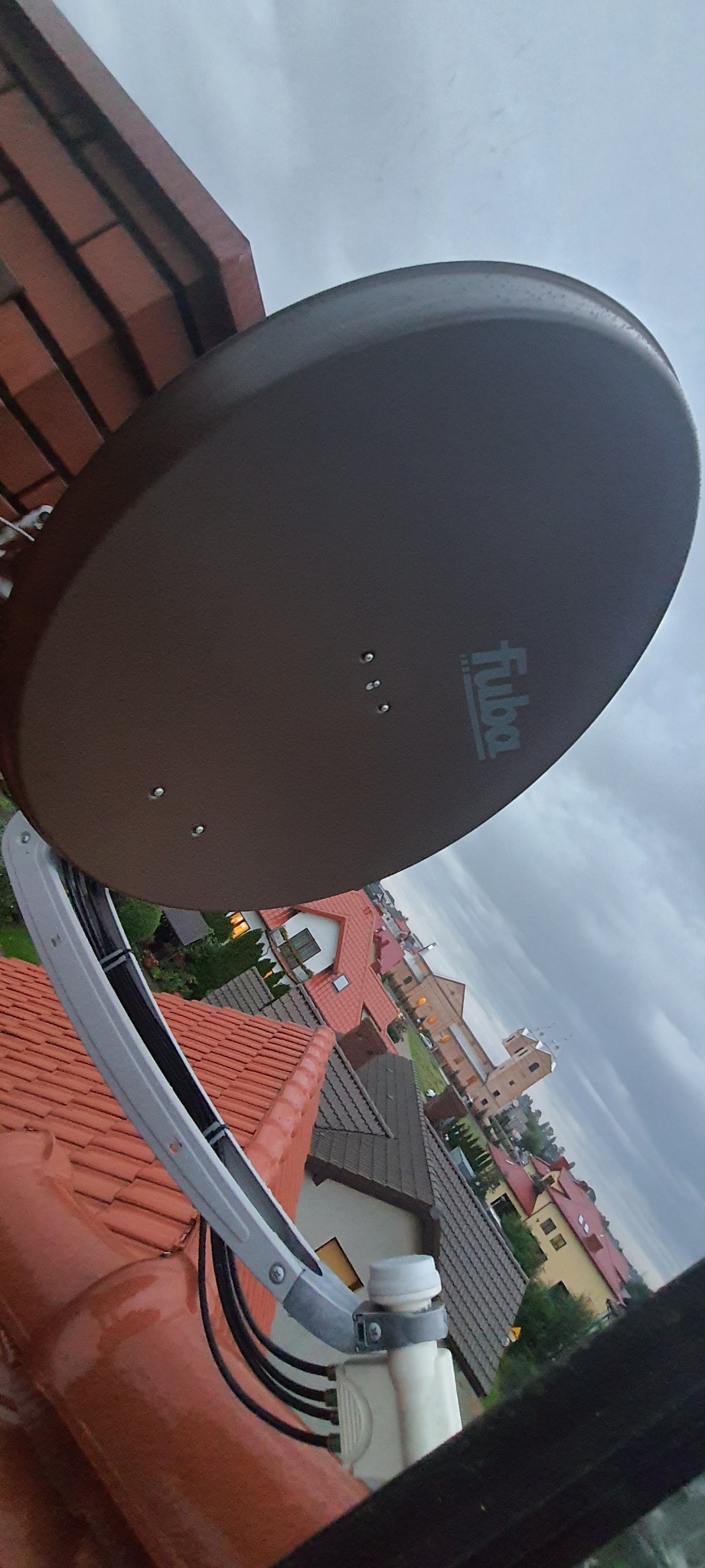 Antena satelitarna marki Fuba zamontowana na pomarańczowym dachu z widokiem na zabudowę miejską i pochmurne niebo.