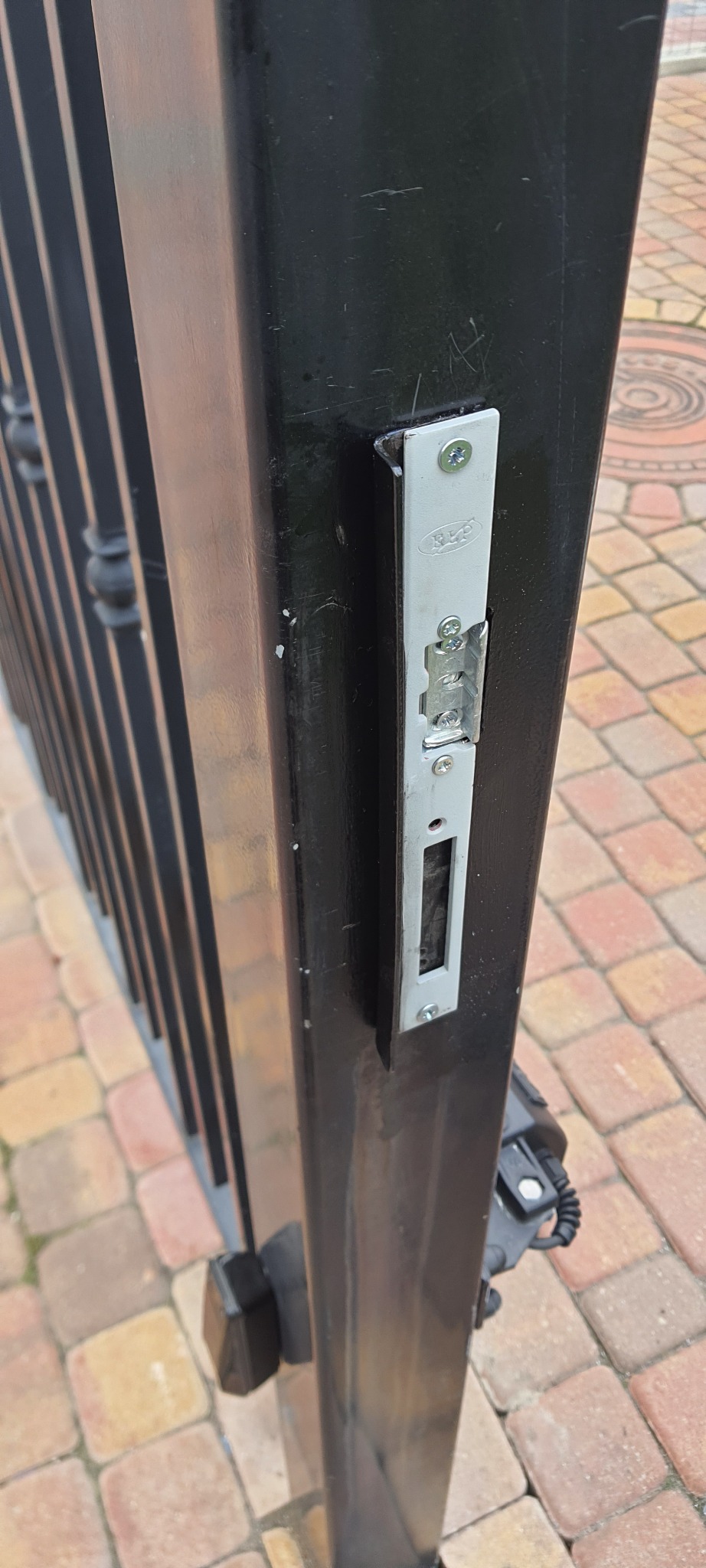 Zamek elektrozaczepowy zamontowany na czarnym słupku bramy wjazdowej z widocznym napędem, na tle kostki brukowej.