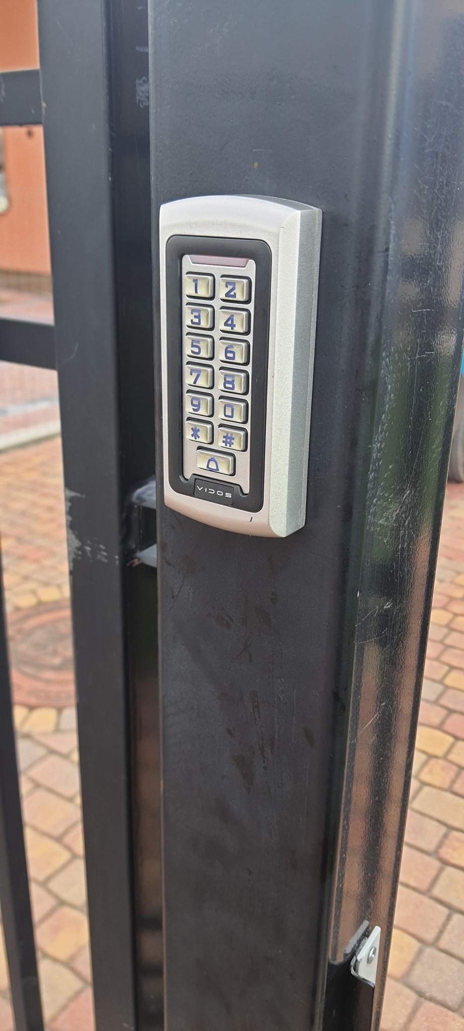 Klawiatura numeryczna domofonu Vidos zamontowana na czarnej metalowej furtce, widoczne ślady użytkowania na powierzchni furtki.