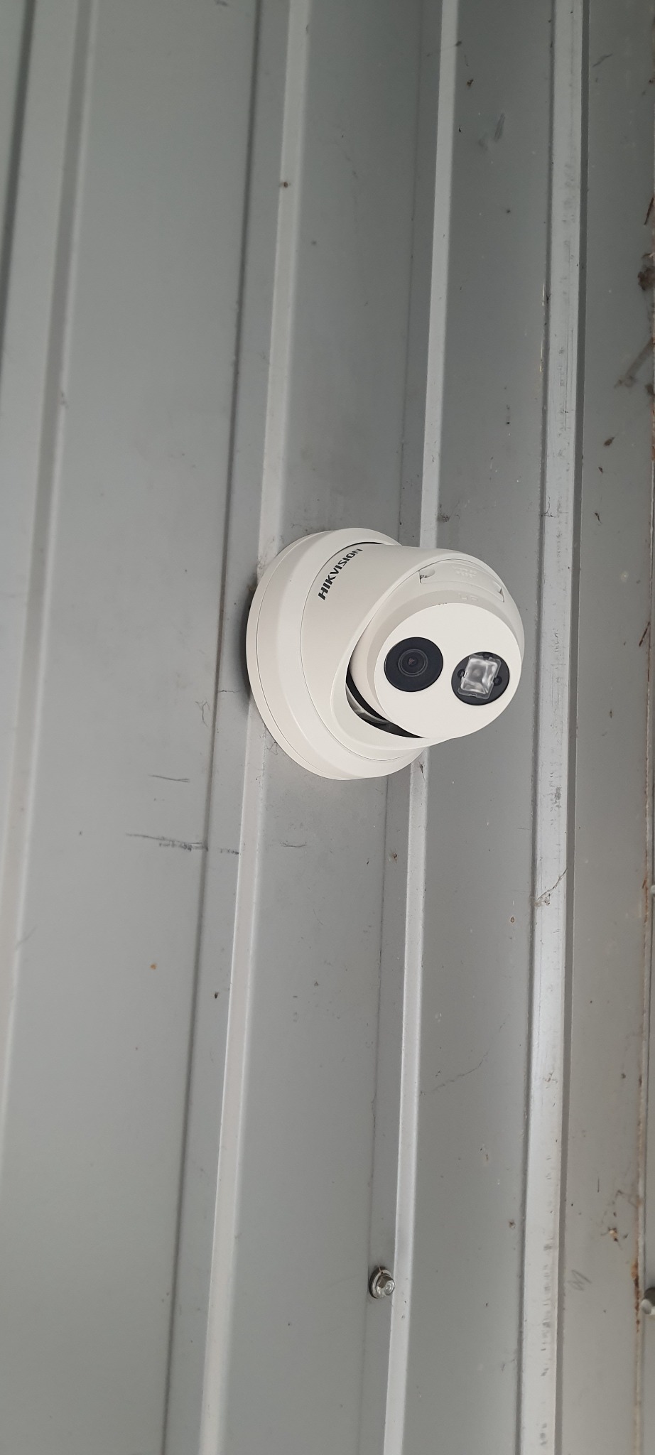 Kamera monitoringu marki Hikvision, zamontowana na szarej, blaszanej ścianie z widocznymi śladami użytkowania i rysami.