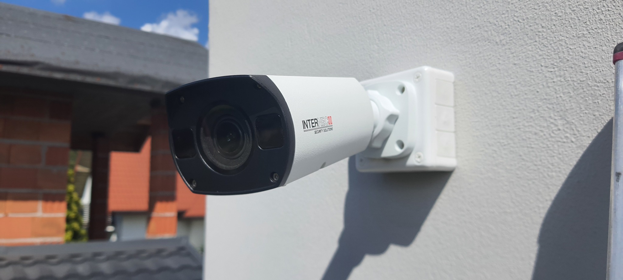 Zewnętrzna kamera monitoringu marki Interneco przymocowana do jasnej ściany budynku z widocznym fragmentem dachu i ceglanej konstrukcji w tle.