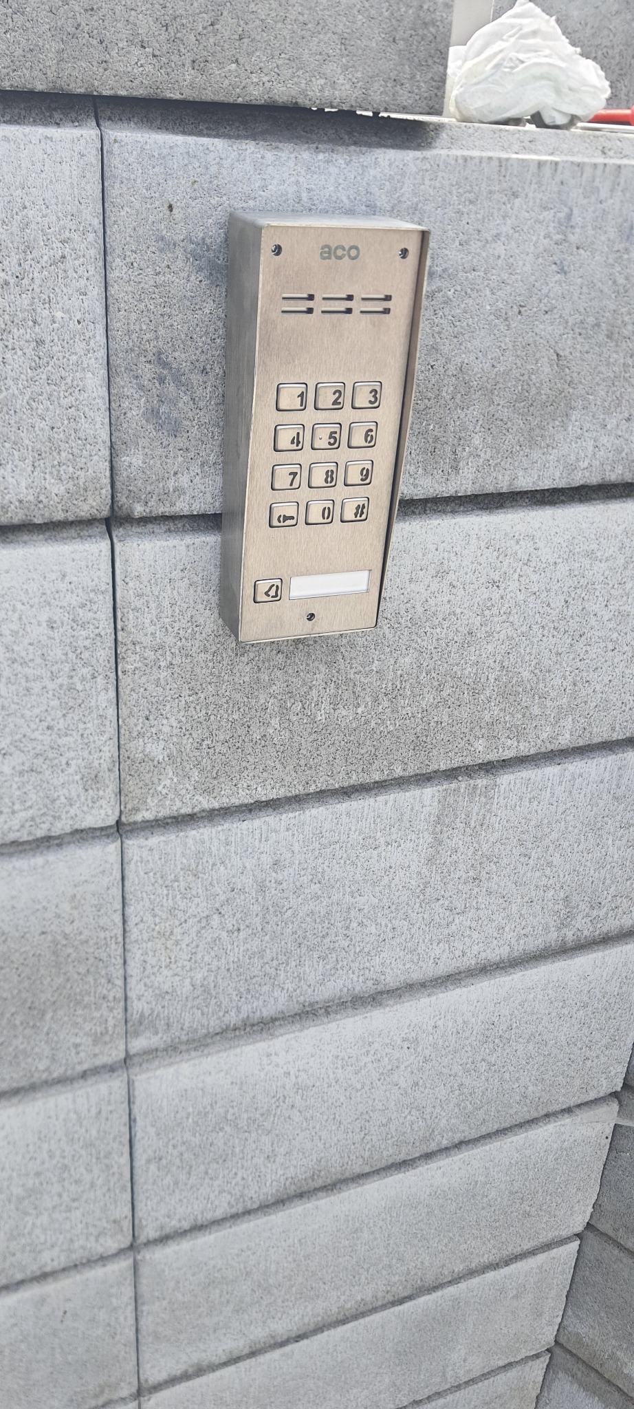 Zewnętrzny panel domofonowy ACO z klawiaturą numeryczną, zamontowany na szarym, betonowym murze.