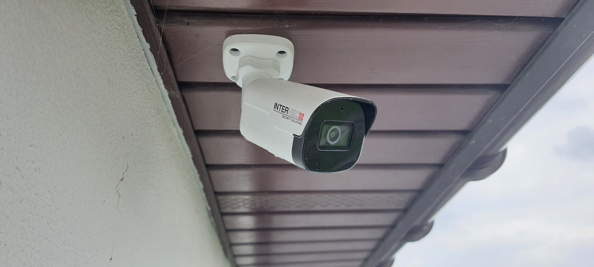 Biała kamera monitoringu Internec Security Solutions zamontowana pod brązowym okapem, widoczne pajęczyny w rogu.