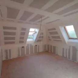 Poddasze w trakcie adaptacji: szpachlowane ściany i sufit z widocznymi łączeniami płyt gipsowo-kartonowych, okna dachowe w skosach, przygotowane do dalszych prac wykończeniowych.