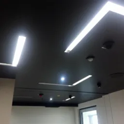 Czarny sufit podwieszany z liniowym oświetleniem LED, widoczne czujniki dymu i kamery monitoringu.