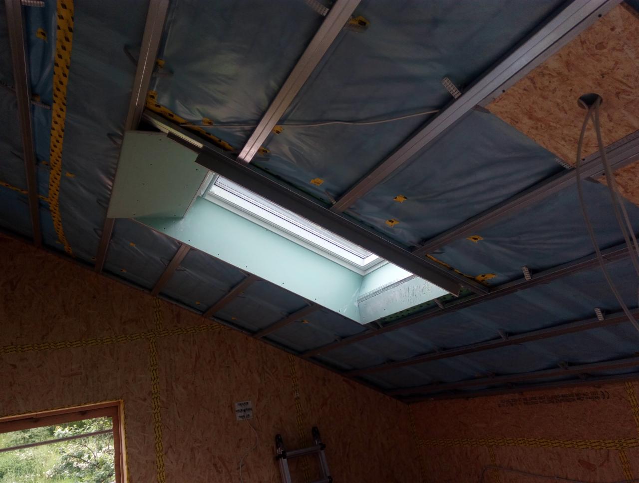 Montaż sufitu: widok na konstrukcję metalową, folię paroizolacyjną i częściowo zamontowaną płytę z otworem na okno dachowe w poddaszu.