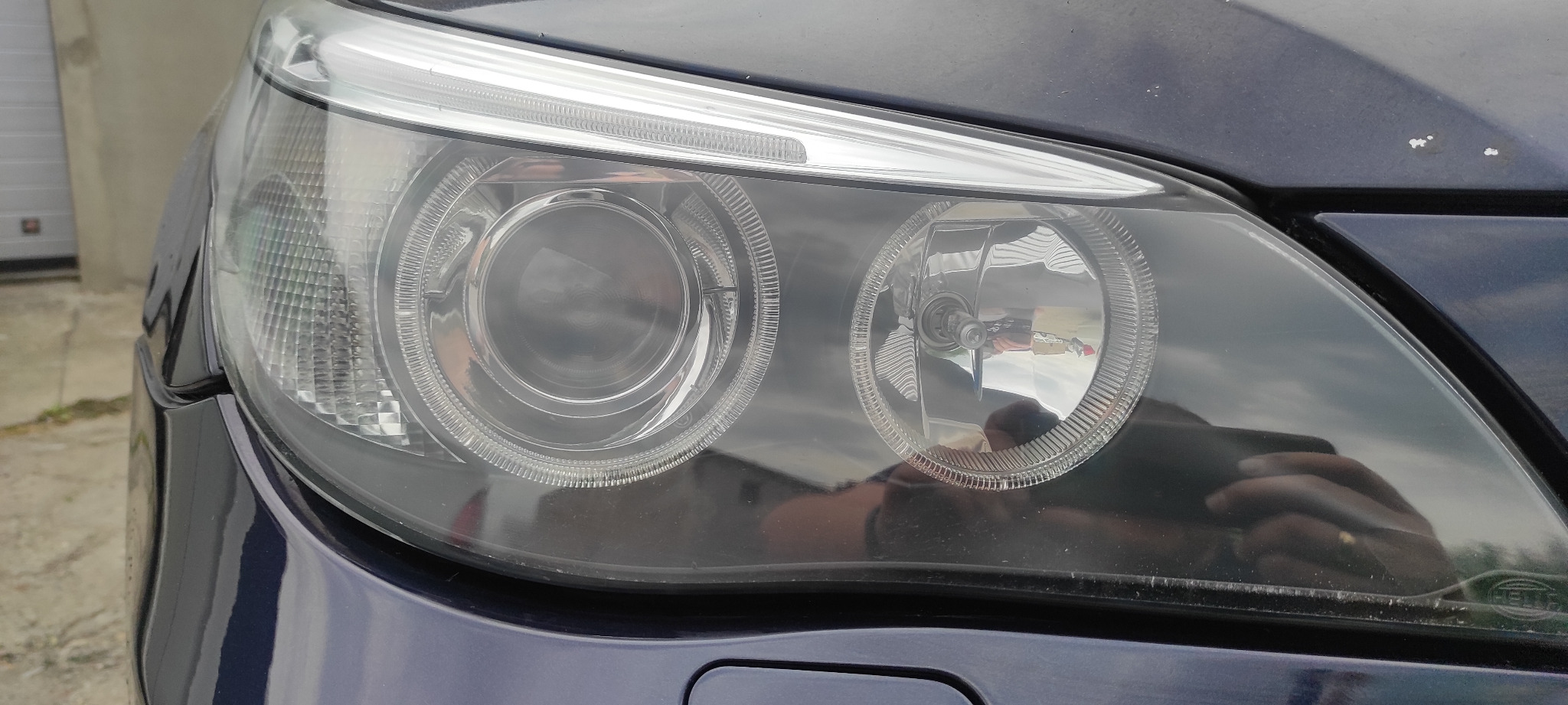 Reflektor samochodowy z widocznym odbiciem osoby robiącej zdjęcie, lekko zabrudzony klosz.