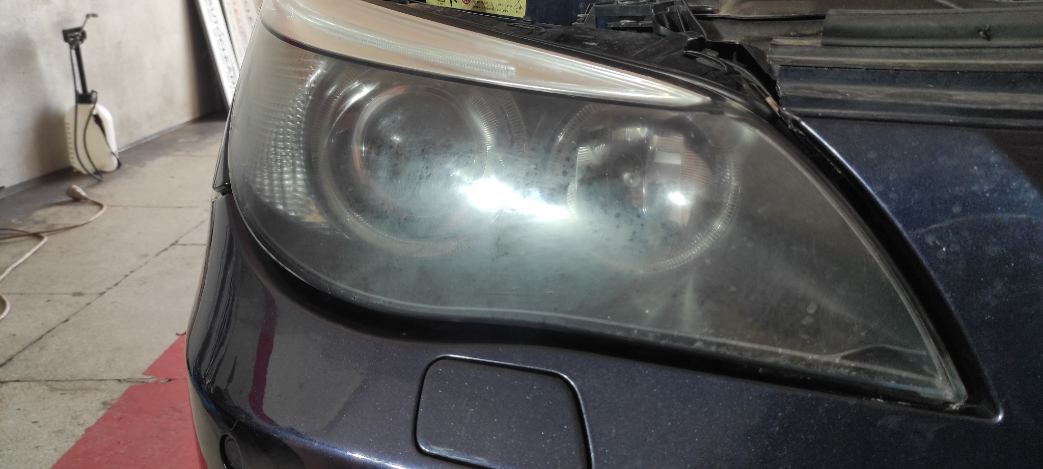 Przednia lampa samochodu z widocznymi śladami zabrudzeń i zmatowienia, wymagająca odświeżenia, zarysowania na kloszu, widoczny fragment zderzaka w kolorze ciemnego granatu.