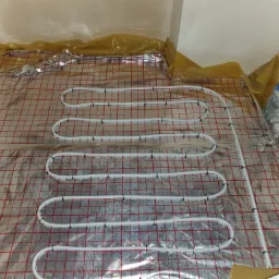 Rozłożona instalacja ogrzewania podłogowego na folii z siatką w pomieszczeniu w trakcie remontu, widoczne rury grzewcze przymocowane do siatki, folia zabezpieczająca ściany.