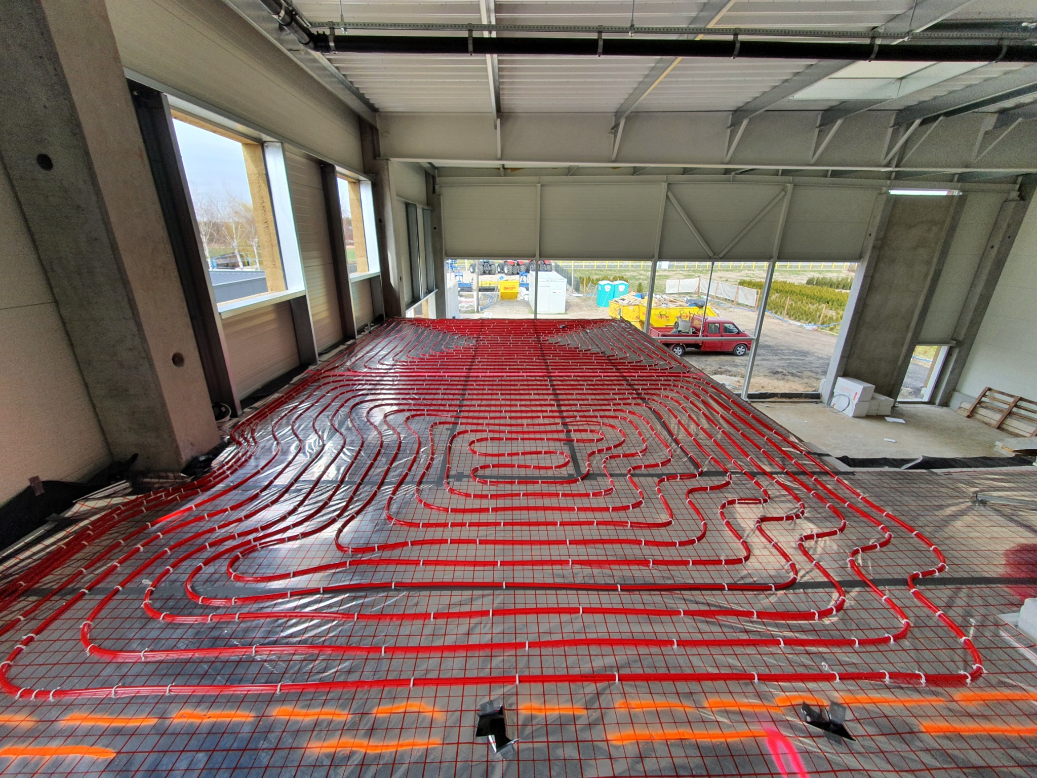 Czerwone rury ogrzewania podłogowego ułożone na siatce w surowym wnętrzu hali, widok z góry na instalację przed wylewką, w tle okna i wyjście na zewnątrz.