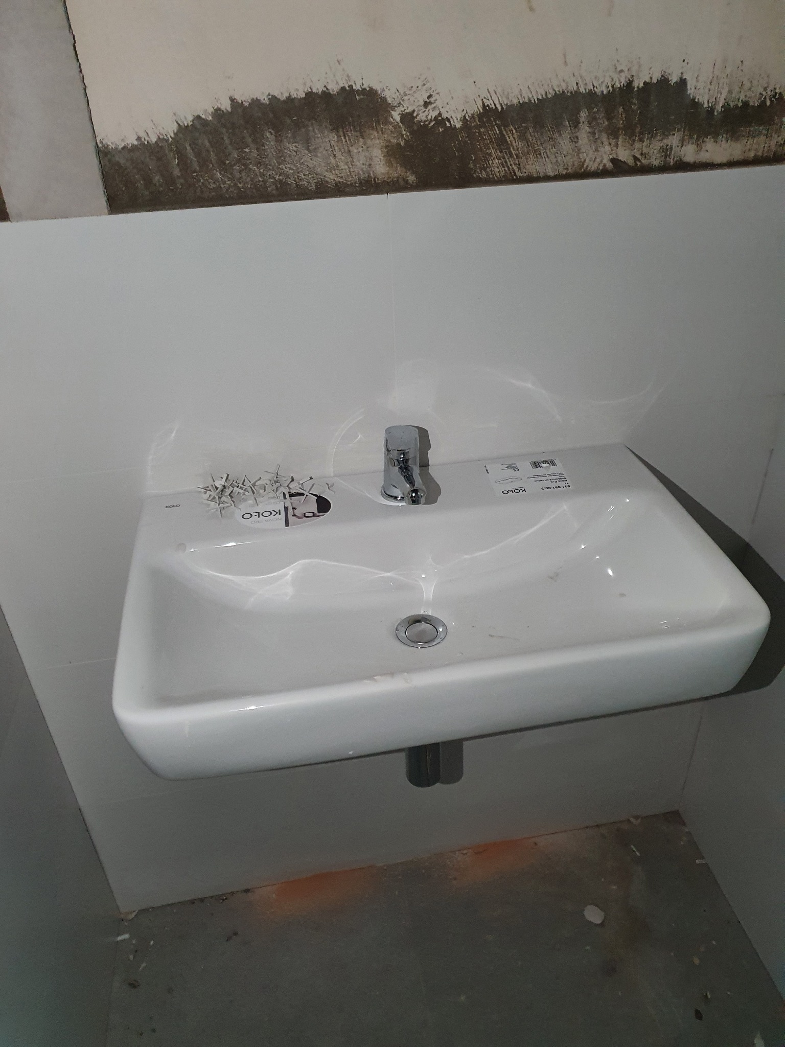 Biała umywalka marki Koło zamontowana na ścianie z chromowaną baterią, widoczne niedokończone wykończenie ściany nad umywalką i elementy montażowe na jej powierzchni.