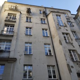 Agencja Alpinistyczna - Byle Wyżej. Damian Postek - Dwóch alpinistów na linach podczas renowacji elewacji kamienicy z balkonami i klimatyzacją, widok z dołu na tle błękitnego nieba.