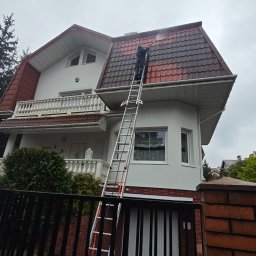 Agencja Alpinistyczna - Byle Wyżej. Damian Postek - Pracownik na drabinie czyści ciśnieniowo dach dwupiętrowego domu z czerwonej dachówki. Widoczne ogrodzenie posesji i fragment elewacji. Pochmurne niebo.