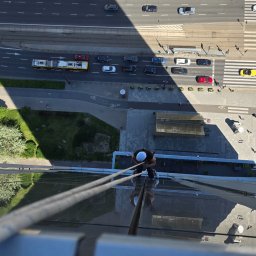 Agencja Alpinistyczna - Byle Wyżej. Damian Postek - Pracownik na wysokości myje szklaną elewację biurowca, widok z góry na ulicę z ruchem miejskim. Bezpieczeństwo zapewniają liny i kask.