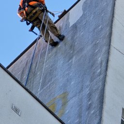 Agencja Alpinistyczna - Byle Wyżej. Damian Postek - Pracownik w uprzęży, na linie, maluje elewację budynku. Widoczne fragmenty pomalowanej ściany i resztki graffiti. Ujęcie z dołu, na tle błękitnego nieba.