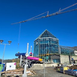 Agencja Alpinistyczna - Byle Wyżej. Damian Postek - Montaż konstrukcji aquaparku w Warszawie z użyciem dźwigu. Widoczne elementy zjeżdżalni i szklana konstrukcja budynku w słoneczny dzień.