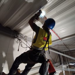 Agencja Alpinistyczna - Byle Wyżej. Damian Postek - Pracownik w uprzęży, w kasku, wierci w suficie hali. Widoczne liny, narzędzia i oświetlenie. Ujęcie z dołu, podkreślające wysokość i specyfikę pracy.