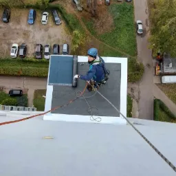Pracownik w uprzęży, z widokiem z góry, instaluje papę na dachu budynku wielorodzinnego; w tle drzewa i zaparkowane samochody.