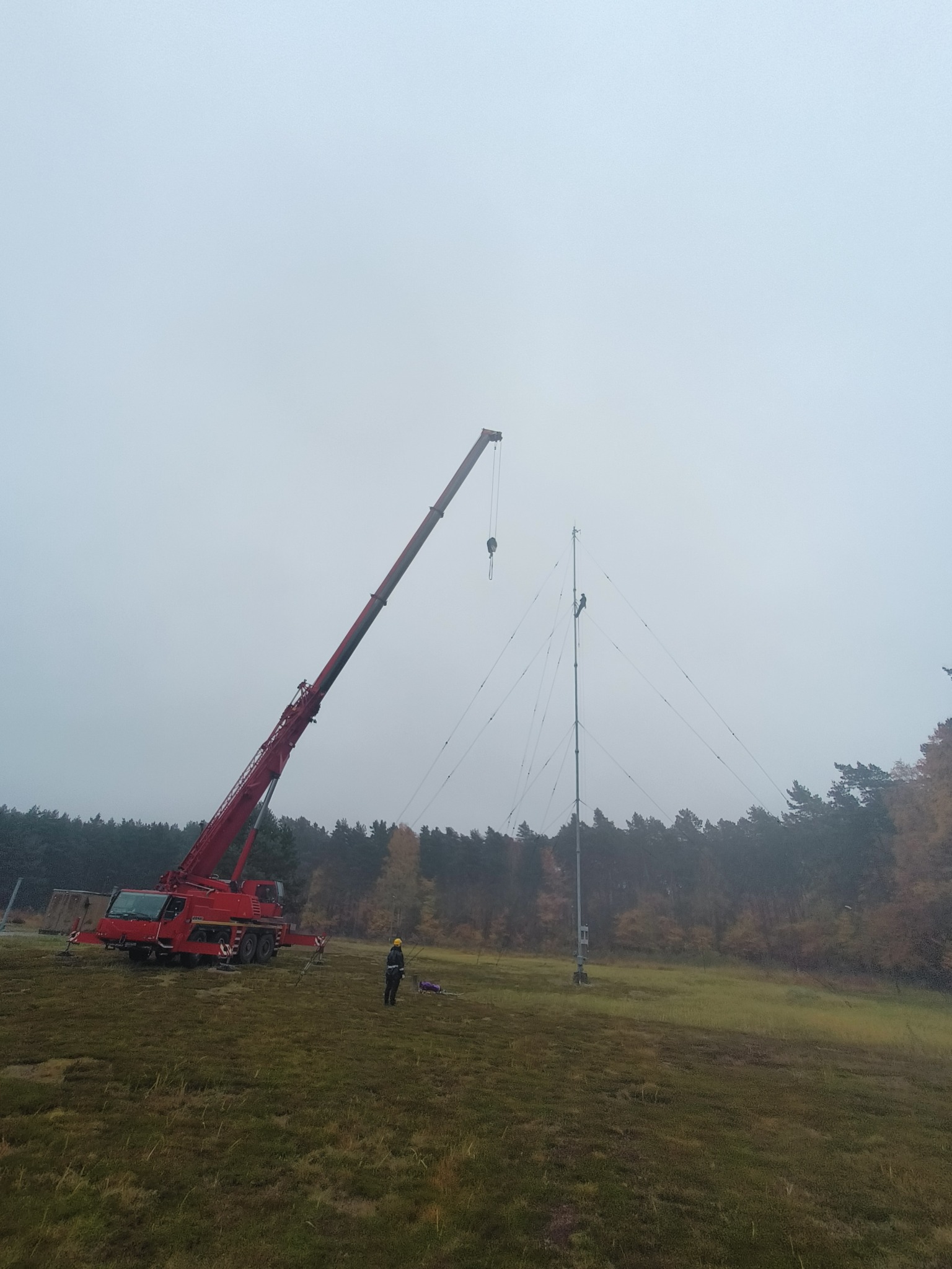 Montaż masztu telekomunikacyjnego przy użyciu czerwonego dźwigu, pracownik w żółtym kasku nadzoruje z ziemi, a drugi wspina się na konstrukcję w pochmurny dzień.