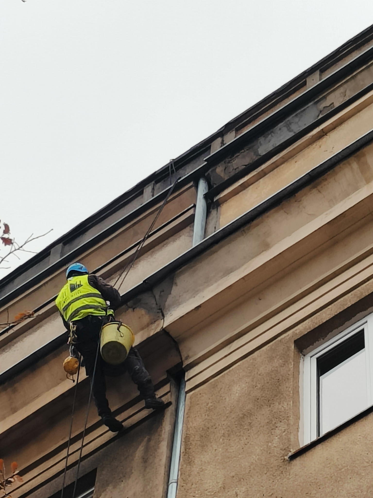 Pracownik na linie, w kasku i żółtej kamizelce, zwisa wzdłuż elewacji budynku z wiadrem, prawdopodobnie podczas prac konserwacyjnych rynny.