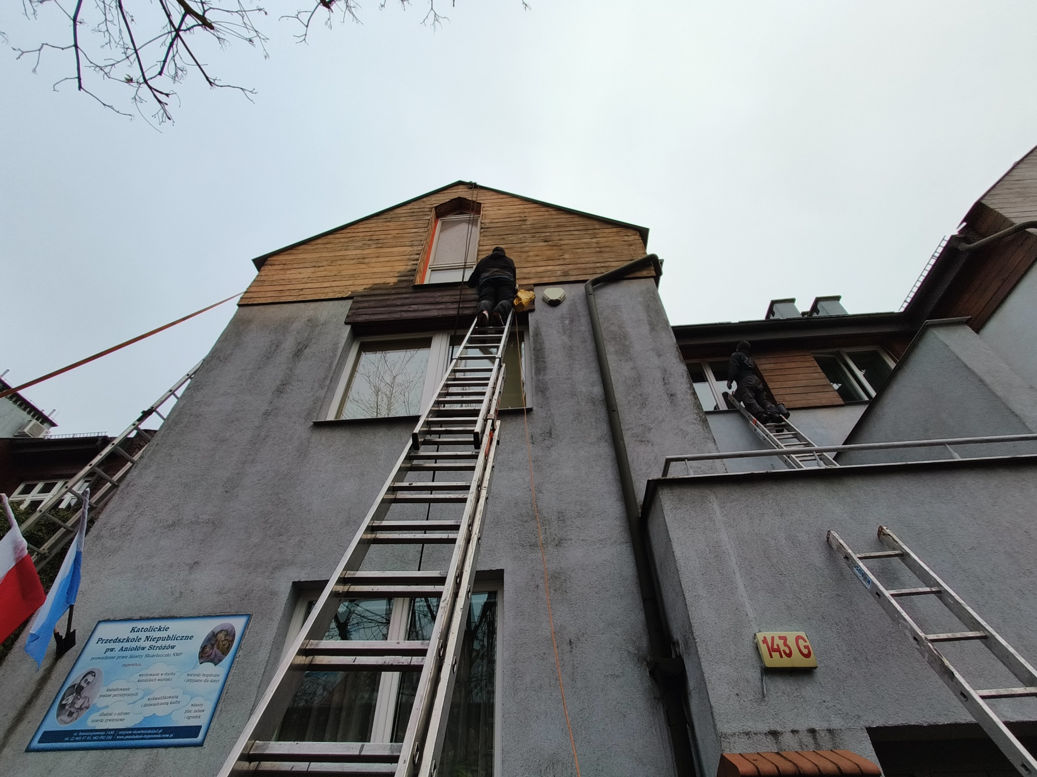 Pracownik na drabinie przy budynku przedszkola, prawdopodobnie wykonujący prace konserwacyjne elewacji lub dachu, widok z dołu na szarą elewację i drewniane elementy dachu, drabina oparta o okno.